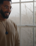 Man wearing EEMedallion Hawk Eye and a beige sweater standing in front of a window