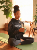 Person sitting on a yoga mat wearing EEHoodie while holding a mug in a home setting with plants and furniture.