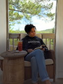 Person sitting on a chair wearing EEHoodie by a window with a drink and book, surrounded by greenery outside.